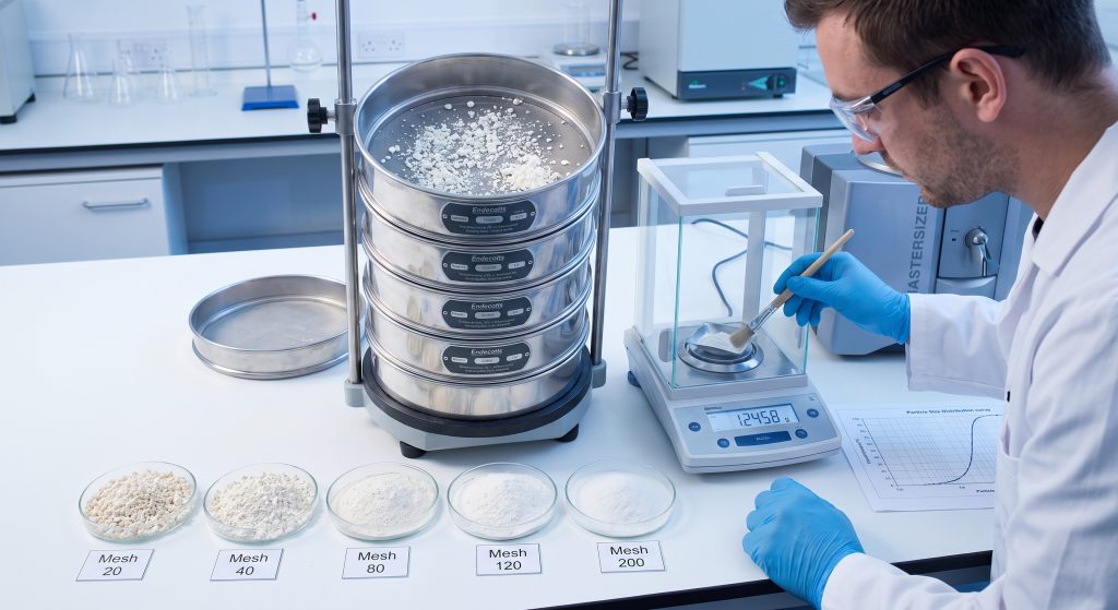 Laboratory sieve stack with graded cassava flour particle samples in different mesh sizes beside particle size distribution chart and QA technician performing granulometry analysis in food testing lab